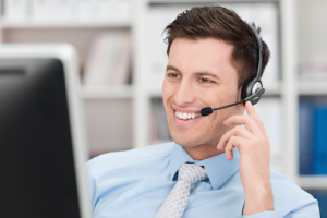 Handsome customer service assistant talking to a customer on the telephone via a headset with computer in the background. Supporting buyers and purchasers